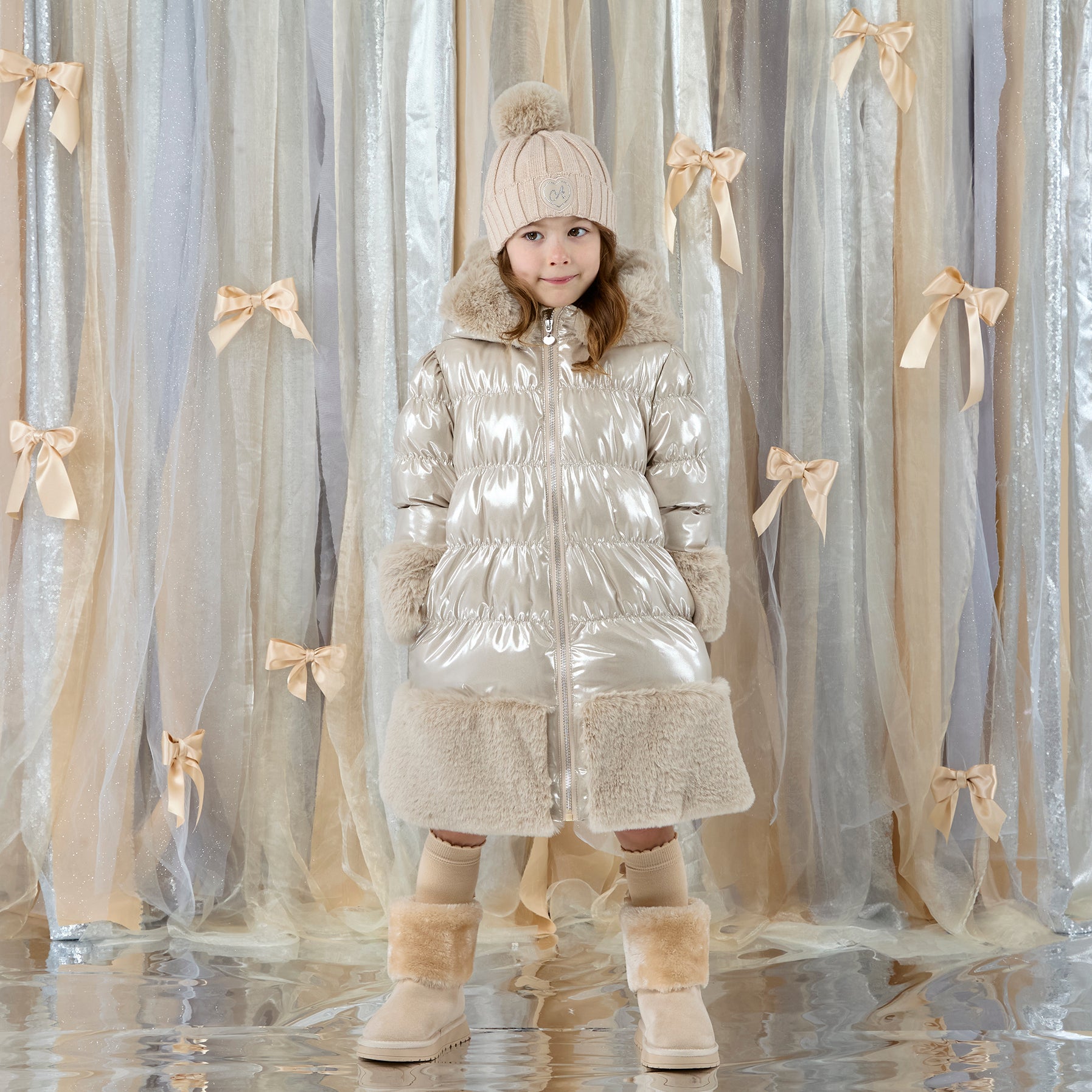 ADEE Eau-de Adee Collection. Introducing the Aurora Jacket, a beautiful fit-and-flare style, finished with soft faux fur trims at the hood, cuffs, and hem. The cosy fleece lining and hood keep you warm, while signature details include a branded badge, logo zip pull, and sweet bows on each arm. Stone Beige