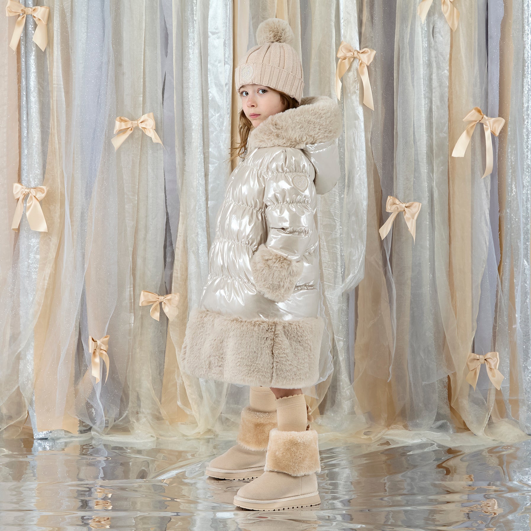 ADEE Eau-de Adee Collection. Introducing the Aurora Jacket, a beautiful fit-and-flare style, finished with soft faux fur trims at the hood, cuffs, and hem. The cosy fleece lining and hood keep you warm, while signature details include a branded badge, logo zip pull, and sweet bows on each arm. Stone Beige