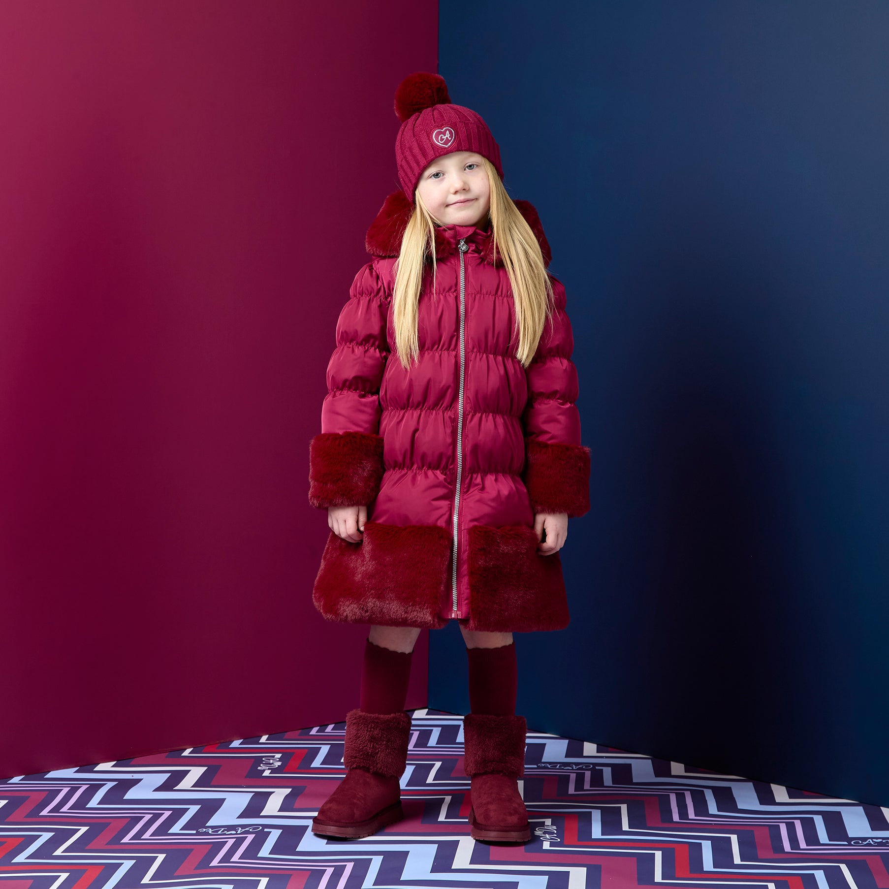 ADEE Chevron Charm theme. Introducing the Aurora Jacket, a beautiful fit-and-flare style, finished with soft faux fur trims at the hood, cuffs, and hem. The cosy fleece lining and hood keep you warm, while signature details include a branded badge, logo zip pull, and sweet bows on each arm. Deep Cherry
