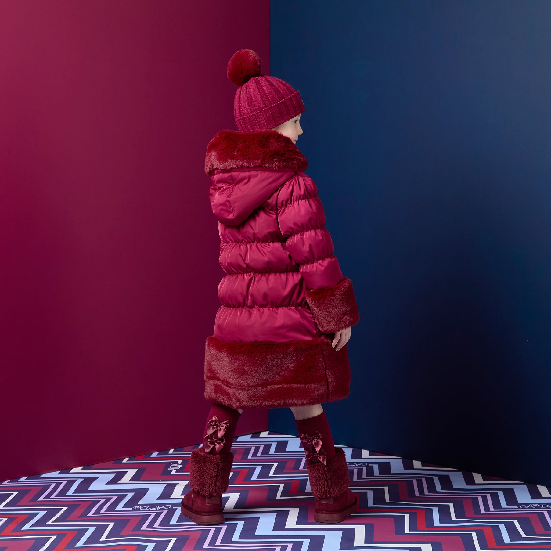 ADEE Chevron Charm theme. Introducing the Aurora Jacket, a beautiful fit-and-flare style, finished with soft faux fur trims at the hood, cuffs, and hem. The cosy fleece lining and hood keep you warm, while signature details include a branded badge, logo zip pull, and sweet bows on each arm. Deep Cherry