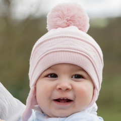 EMILE et ROSE Griffin - Baby pink knitted helmet-shaped hat with ear flaps and small pompoms trimming the ends of the ties below each ear. The main feature is the fun, over-size pompom on the top!