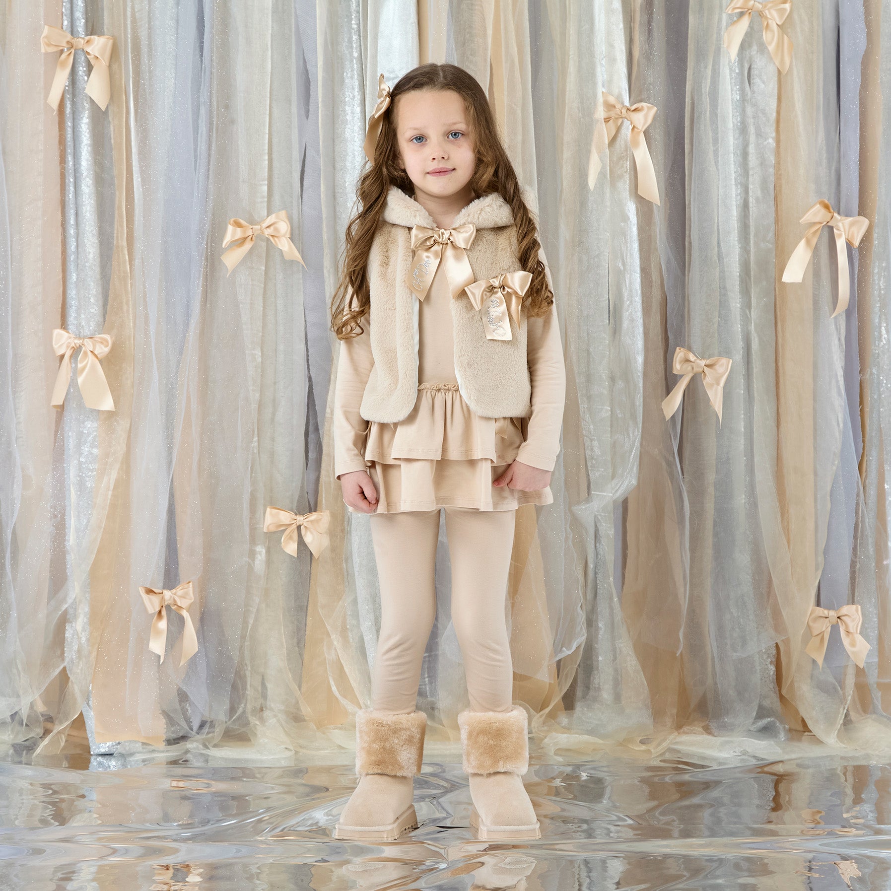 ADEE Eau-de Adee Collection. The Anna gilet come in fabulous faux fur and is finished with a beautiful appliqué bow on the chest. Stone Beige